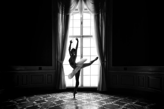 A black and white photograph of a ballerina in a tutu performing a graceful arabesque pose in front of a large bright window in an elegant room.