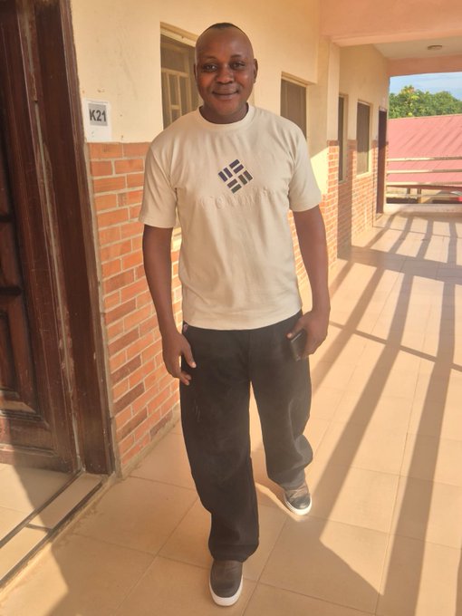 A smiling man wearing a beige t-shirt with a black logo and dark pants stands on a tiled walkway next to a brick wall, holding a phone, with a sign reading "K21" on the wall behind him.