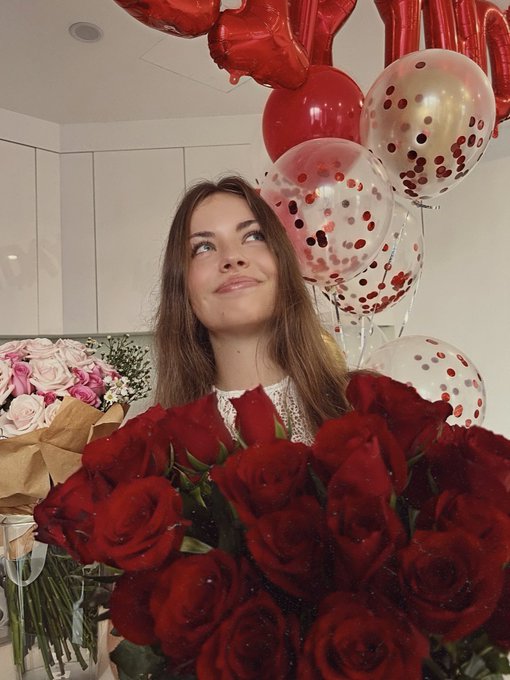 A young woman smiles while holding a large bouquet of red roses, with heart-shaped balloons and '24' decorations behind her.