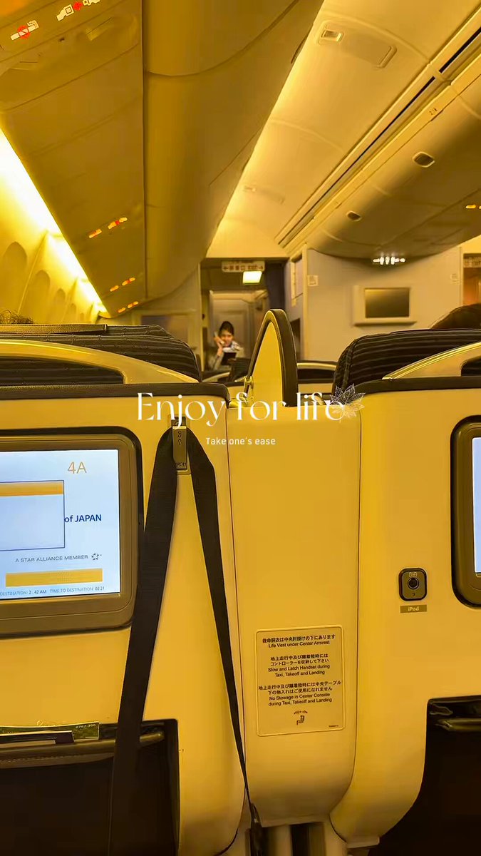 The image shows a person sitting inside an airplane, with their back facing the camera. They are seated between two windows that have yellow seats. The background of the photo features a sign that reads "Enjoy life", which seems to be related to the airplane experience or the airline itself.