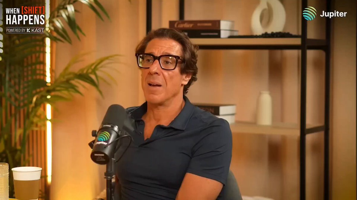 The image features a man wearing glasses sitting at a desk with a microphone, suggesting that he might be hosting a podcast or participating in an interview. The background includes a bookshelf filled with various items such as vases and books, indicating that this could be a home office or a studio setting. The post also mentions Bitcoin and XRP, which are digital currencies that have been the subject of much discussion and speculation due to their potential impact on the financial landscape.