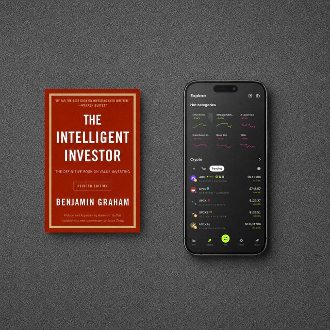A copy of Benjamin Graham's "The Intelligent Investor" rests beside a smartphone displaying the Robinhood crypto trading app, with trending tokens like ARB, SPY, SPCA, SPC20, and bitcoin visible—visually bridging Graham's classic value investing principles with modern onchain markets.