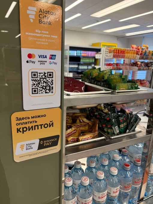 A close-up of a checkout counter displays an Alatau City Bank sign accepting crypto payments via Binance Pay (with a QR code and supported payment options including Visa, Mastercard, Apple Pay, Samsung Pay, Google Pay), with snack shelves and bottled water visible in the background—illustrating Binance Pay's real-world integration across 5,000 POS terminals in Kazakhstan.