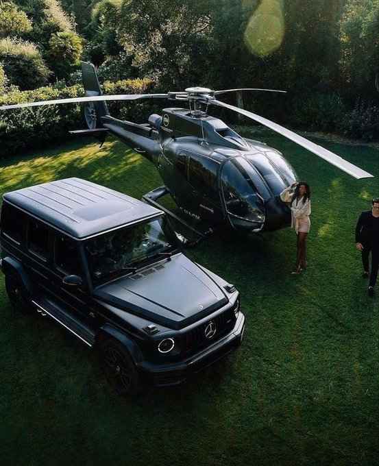 Good morning from the lap of luxury — a sleek black Mercedes G-Wagon parked beside a matching helicopter on a lush green lawn, with a stylishly dressed woman ready for a sky-high start to the day.