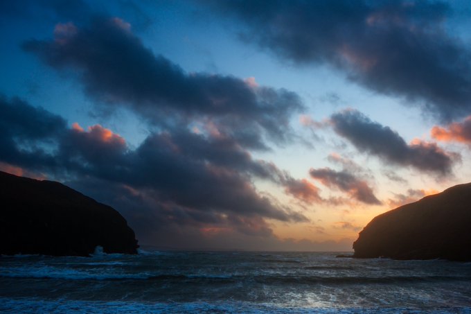 A serene coastal landscape at sunset showing dark cliff silhouettes framing a view of the ocean with dramatic clouds tinted pink and orange in the sky.