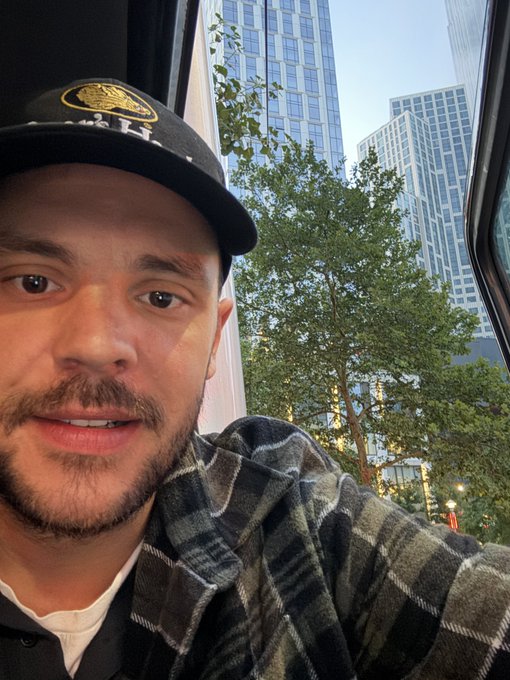 A selfie of Justin Bieber wearing a black baseball cap and a gray plaid flannel shirt, taken near a window with a view of city skyscrapers and trees.