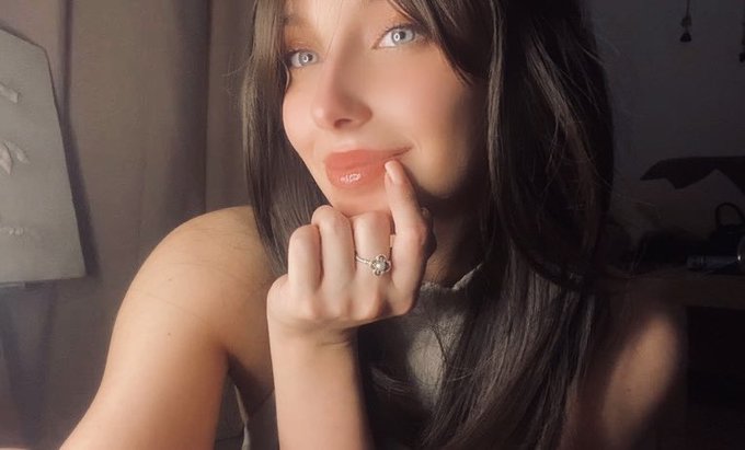 A close-up selfie-style photograph of a young woman with long dark hair and blue eyes, wearing lip gloss and a ring, with her finger near her lips.