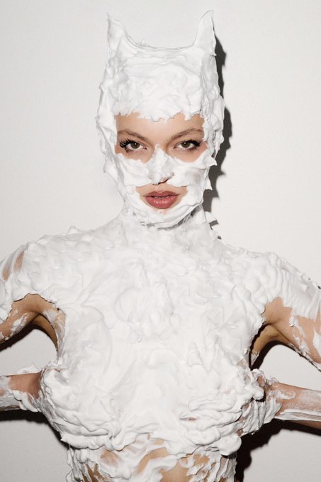 A fashion editorial photograph of a woman wearing an avant-garde white textured bodysuit and headpiece with cutouts for her eyes and mouth, set against a plain white background.