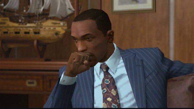 A man in a pinstripe suit and floral tie sits with his hand over his mouth in an office setting, featuring a model ship in the background.
