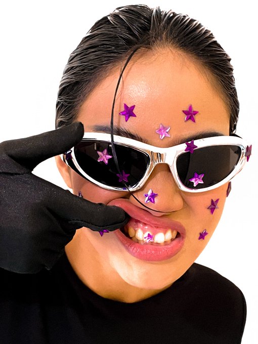 A person with slicked-back wet hair wearing white sunglasses, a black glove, and pink star face stickers makes a playful expression by pulling down their eyelid.