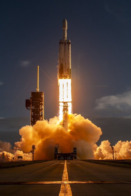 A rocket launches from the launchpad, billowing bright orange exhaust plumes into the dusk sky, with the launch tower standing to the side as flames illuminate the runway below. The image evokes the spirit of reaching for ambitious goals, fitting the optimistic tone of the post about MarsCoin and forward-thinking friendship with time.