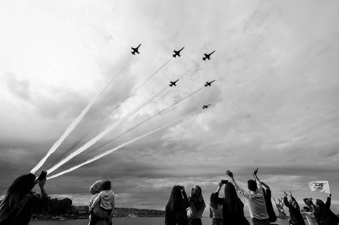 A black and white photograph of an air show with six fighter jets flying in formation leaving smoke trails, while a crowd of spectators watches from below with raised arms and cameras.