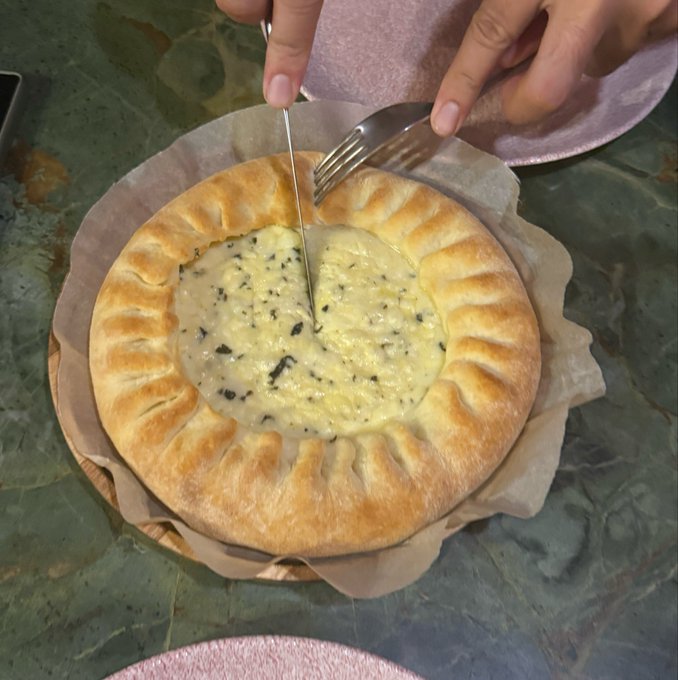 A person uses a knife and fork to cut into a round pastry pie filled with a creamy custard containing dark specks, served on parchment paper.