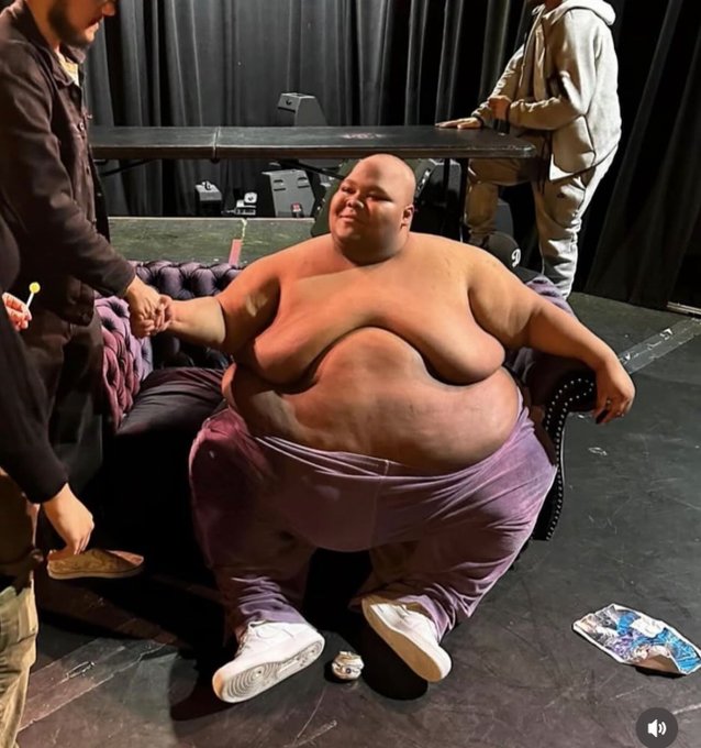 A very large, shirtless man sits on a small couch on a stage, holding hands with someone standing beside him, while another person stands behind holding a DJ controller. The image humorously visualizes "fat bags" by depicting an extremely heavy-set person as a hyperbolic representation of oversized crypto holdings ahead of a bull run.