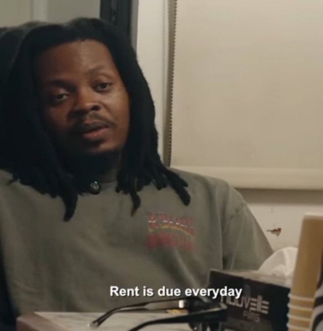 A man with long dreadlocks wearing a green t-shirt sits indoors. The text overlay reads 'Rent is due everyday' and a 'TELEVISIONS' remote is visible in the foreground.