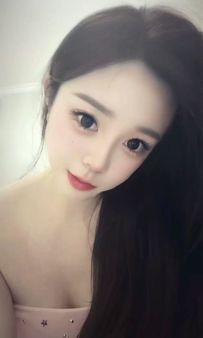 A close-up selfie of a young woman with long dark hair, wearing a pink strapless top, looking at the camera with a slight smile.