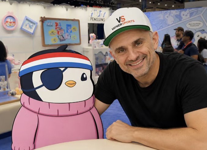 A man wearing a 'VaynerSports' cap smiling alongside a cartoon penguin mascot wearing an eye patch and pink sweater, at what appears to be a 'Study Penguins' booth.