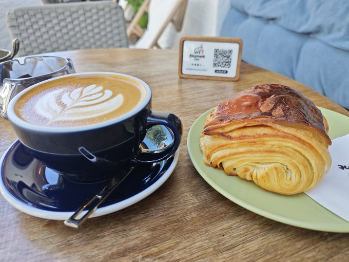 A café table scene featuring a cup of latte with beautiful leaf-shaped latte art alongside a flaky croissant pastry on a green plate, with a QR code sign in the background.