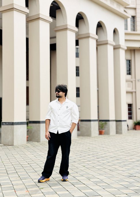 A man with dark hair, a beard, and sunglasses stands in front of an arched colonnade, wearing a white button-down shirt and loose black pants.