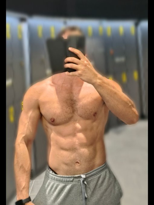 A shirtless, muscular man taking a mirror selfie in a gym locker room while wearing gray sweatpants.
