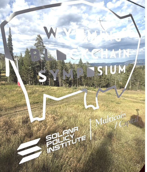 The image shows a frosted glass window etched with the "Wyoming Blockchain Symposium" logo, set against a backdrop of a grassy field, pine trees, and mountains. At the bottom are logos for the Solana Policy Institute and Multicoin Capital, reinforcing the crypto and blockchain industry context of the event referenced in the post about Day 2 of the symposium.