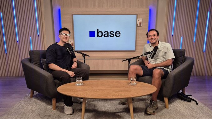 Two men sit in gray armchairs across a round wooden coffee table during a podcast recording, with a large screen behind them displaying the "base" logo. The setup matches the post announcing an upcoming podcast interview with the Base team, featuring advisor Carlos J. Melgar as one of the participants.