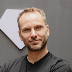 A man with short hair and a stubble beard, wearing a black t-shirt, stands in front of a gray background with a partial white geometric shape visible. The image accompanies the news of Tether CEO Paolo Ardoino's denial of plans to develop a company blockchain.