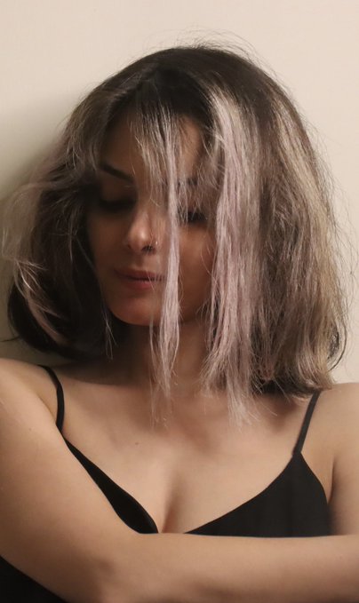 A woman with messy, light purple-streaked hair partially covering her face, wearing a black spaghetti strap top, looking downward. The "me vs. me" caption suggests a contrast between her styled appearance in the image and a more disheveled or bare-faced version, highlighting internal self-comparison.