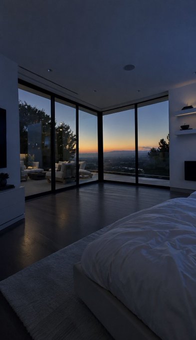 A modern bedroom with floor-to-ceiling windows offers a panoramic view of a city skyline at sunset, featuring dark wood floors, a white bed, and an outdoor patio visible through the glass.