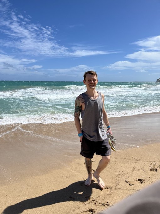 A smiling man with a tattoo on his right arm stands barefoot on a sandy beach, wearing a gray tank top and dark shorts, with ocean waves and a blue sky behind him.