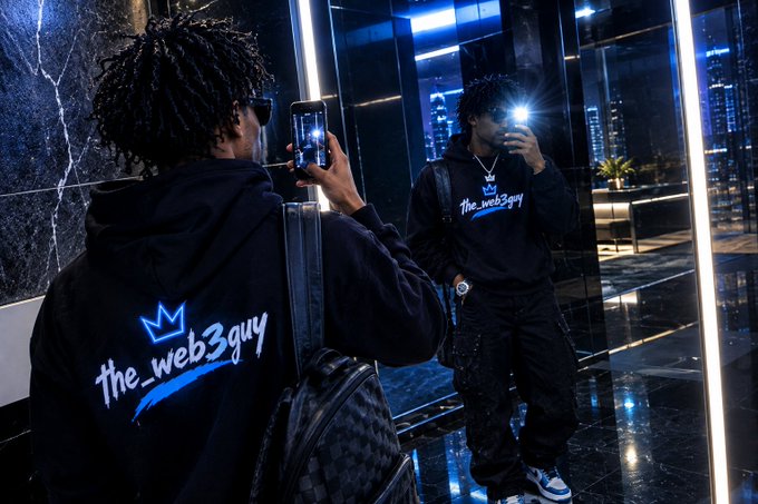 A young man with dreadlocks takes a mirror selfie wearing a black "The_web3guy" hoodie with a crown logo, in a sleek modern lobby at night.
