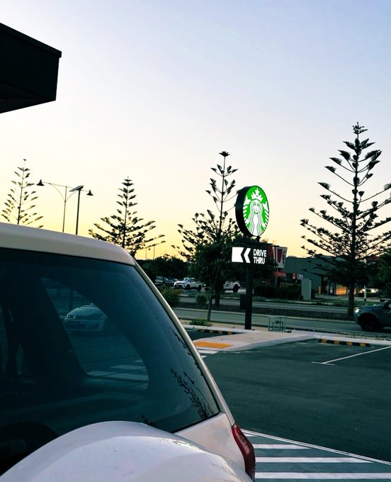 An outdoor view at sunset showing a Starbucks drive-thru sign and parked cars in a lot lined with pine trees.