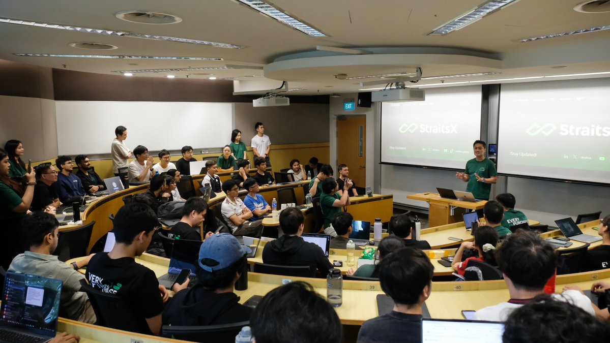 A presenter in a green shirt speaks at a podium in a packed university lecture hall during a hackathon, with two large screens displaying "StraitsX - Stay Updated" alongside social media handles, as attendees with laptops listen attentively from tiered seating.