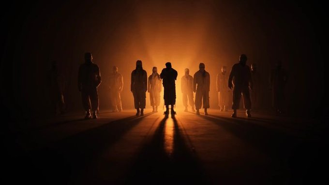 A dramatic silhouette image of multiple hooded figures standing in a circular formation against a glowing orange backlight, with long shadows cast on the ground.