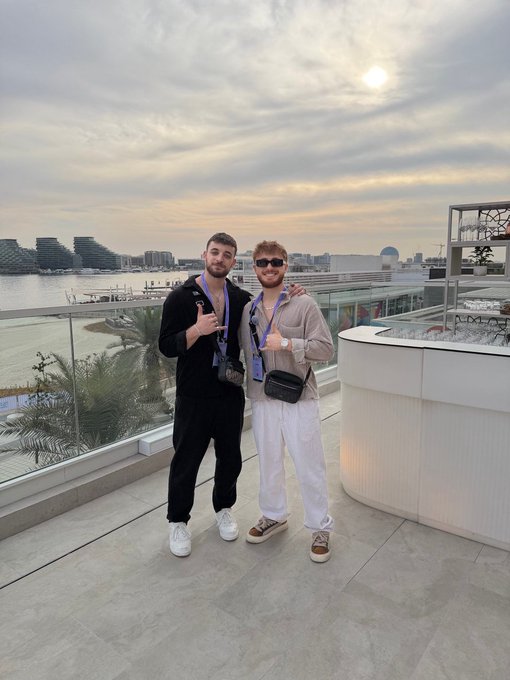 The image shows two men standing next to each other, both wearing black clothing with one of them holding a purple lanyard around his neck. They are posing for a photo together against an outdoor setting featuring palm trees and a building in the background. The caption "GM" is present in the image, which could be related to their location or possibly something else that caught their attention during the photo session.