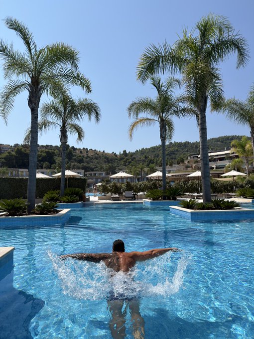 A man splashing water in a luxurious resort pool surrounded by tall palm trees, with hotel buildings and sun loungers visible in the background under a clear blue sky.