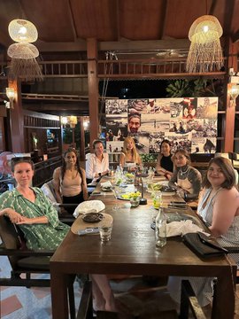 A group of people seated at a long wooden table in a warmly lit restaurant with hanging lamps.