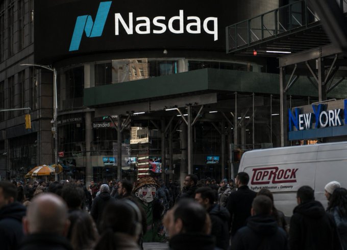 Street-level photograph of the Nasdaq building in New York City with crowds of people on the sidewalk and a 'TOP ROCK' van in the foreground.