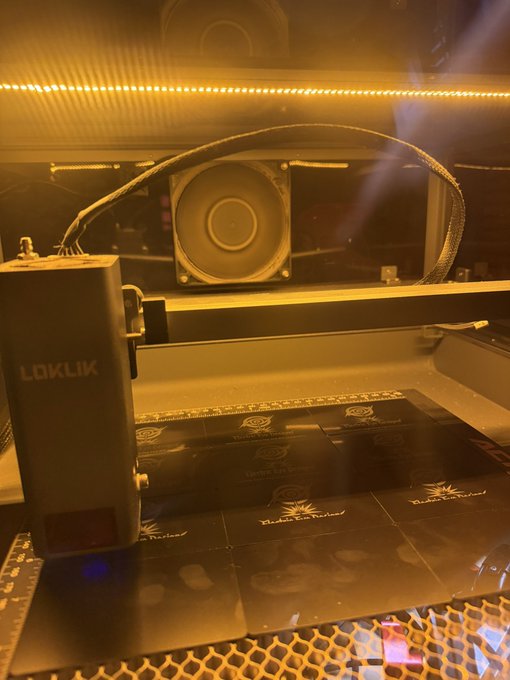 A LOKLiK engraving machine inside an illuminated enclosure, with a speaker visible in the background and engraving sheets on the work surface.