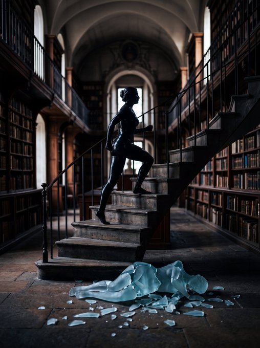 A dark, humanoid figure ascends a staircase in a grand library, leaving behind shattered ice-like fragments at the base.