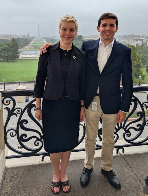 A man and woman pose together on a balcony with a wrought iron railing, with the Washington Monument and the National Mall visible in the background.