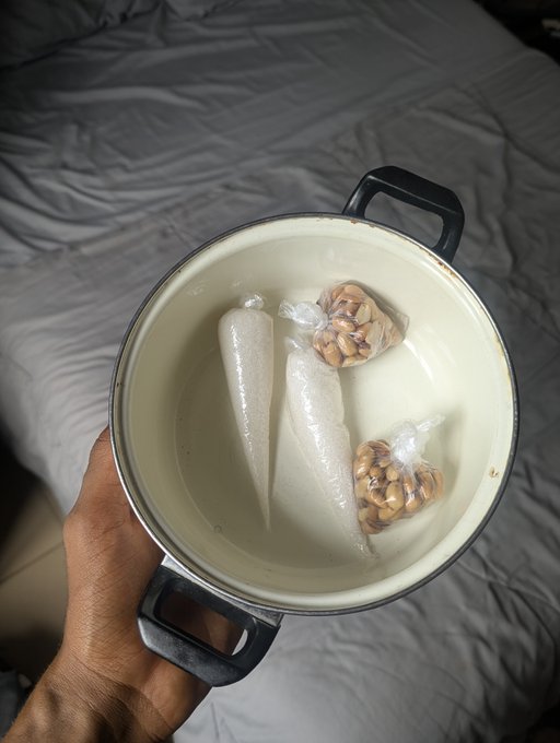 Hand holding a small cooking pot containing two clear soup dumplings or rolls and two small mesh bags of beans or grains in liquid.