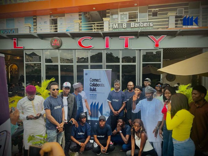 A group of people pose for a photo outside a venue named 'L CITY', with a banner reading 'Connect. Collaborate. Build.' and an 'AKM Abuja Web3 Connect' logo.