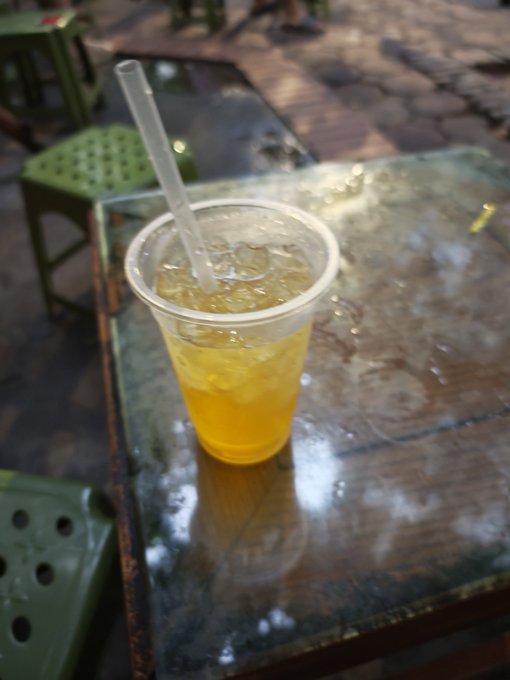 A clear plastic cup filled with a yellow iced beverage and a straw sits on a weathered glass-topped table, with green plastic stools visible in the background.