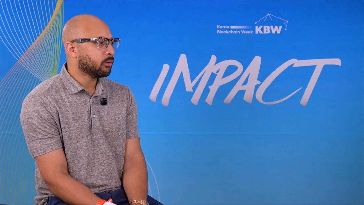 The image features a man wearing glasses sitting down with his hands crossed. The background of the image has a blue color, which appears to be part of an advertisement or promotional material for a conference. The text "KBNB" can also be seen in the image. The main focus is on the man and the blue background, but there are some words related to the event that might provide additional context about the conference.