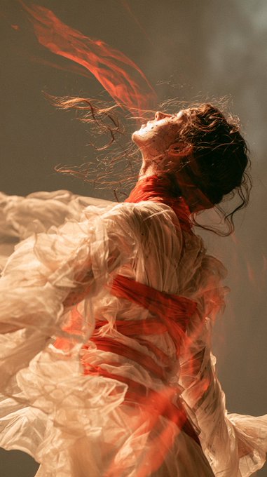 A woman in a flowing white garment tilts her head back dramatically, with red fabric swirling around her in an artistic, ethereal composition.