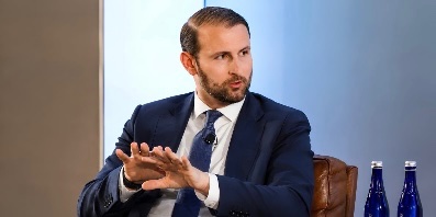 The image shows a clean-cut man in a dark navy suit and tie, seated and gesturing with both hands raised, palms open as if explaining or emphasizing a point during a panel discussion or interview. The caption from the post identifies him as White House advisor Patrick Witt, who is advocating for the CLARITY Act with a vote scheduled for September 15, suggesting that passage could benefit Bitcoin. The man's engaged, expressive hand gestures in the image visually reinforce his role as an advocate passionately making his case for the legislation and its potential impact on the crypto market.