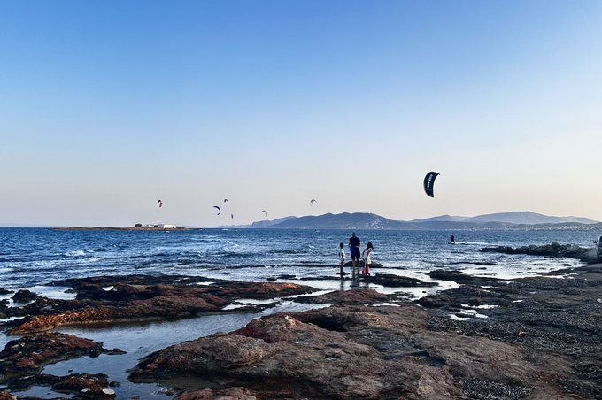 A scenic coastal view with people standing on rocky tide pools watching kitesurfers on the water under a clear blue sky.