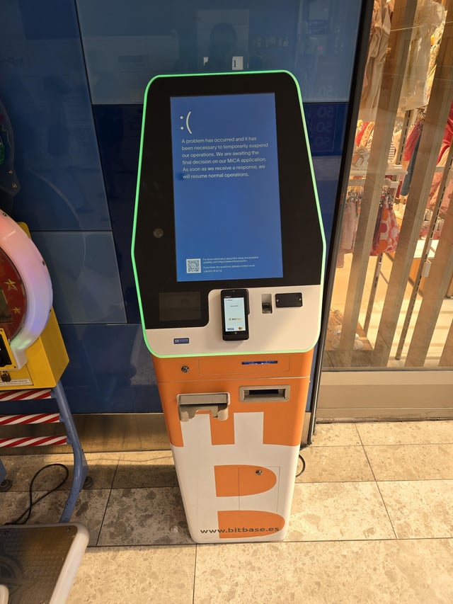 A Bitcoin ATM (BLITBASE) displays a sad face and an error message: 'A problem has occurred and it has been necessary to temporarily suspend our operation. We are awaiting the final decision on our MICA application. As soon as we receive a response, we will resume normal operations.'