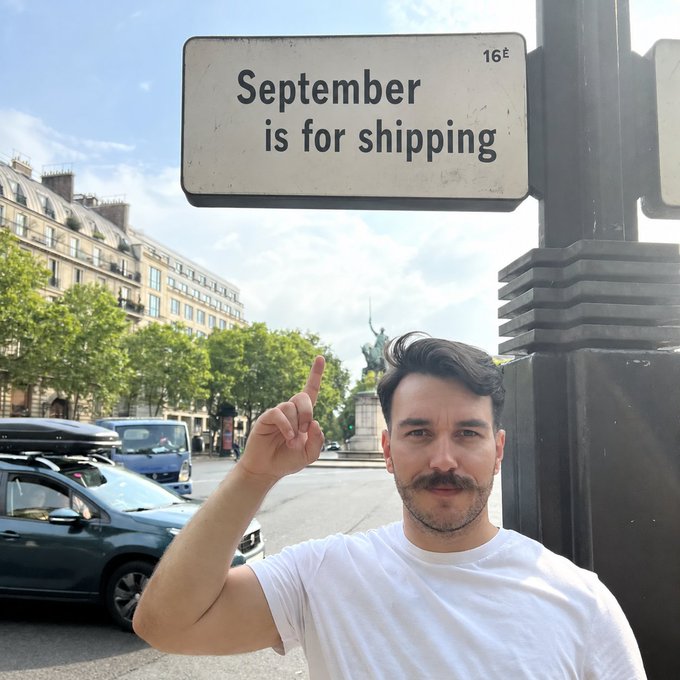 A man with a mustache stands on a Paris street pointing up at a street sign that humorously reads 'September is for shipping.' The Statue of Liberty replica and classic Parisian buildings are visible in the background.